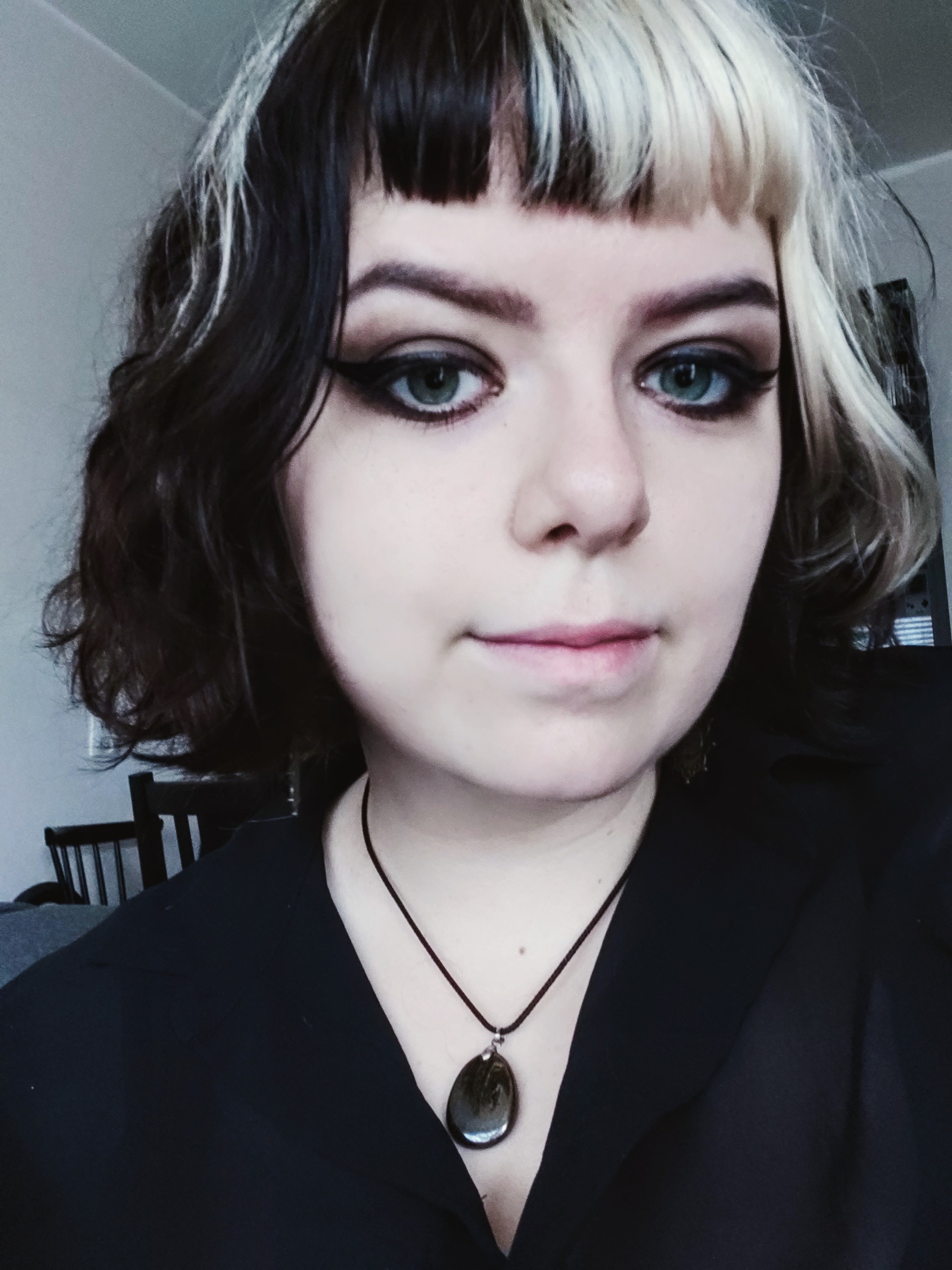 A shoulder's-up selfie style picture of Jade Borra. They have chin-length black hair with white streaks, dark makeup and dark clothing